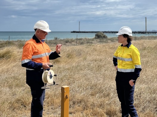 Photo of a man and woman in high visibility jackets and hard hats, with the Cockburn Sound in the background.