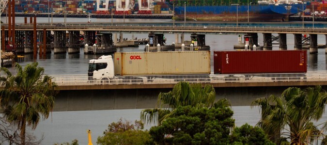 Photo of truck towing two containers over a bridge with a port in the background